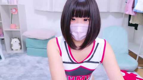 Media: Video of an East Asian woman with straight, dark hair, wearing a white and red dress, pink face mask, and seated on a soft, light blue cushion in a cozy room with white walls, pastel decor, and a small stuffed animal.