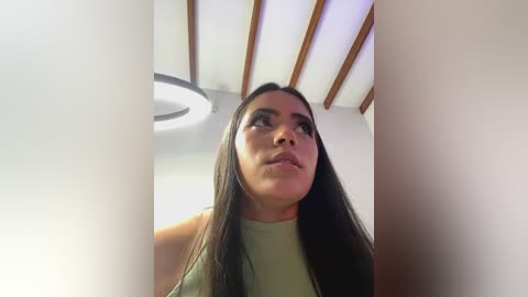 Media: A video of a Latina woman with long, straight, dark hair, wearing a green top, looking up. She stands in a modern, minimalist room with a wooden ceiling and a round light fixture.