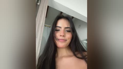 Media: Video of a topless young woman with long black hair, light skin, and a slight smile, standing in a modern bathroom with beige walls and a shower curtain.