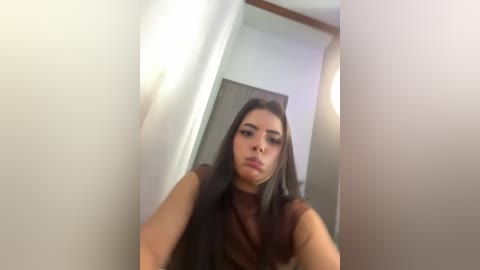 Media: Video of a young woman with long, dark hair and light skin, wearing a brown top, standing in a doorway with white walls and a wooden door in the background.