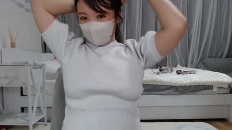 Media: Video of a woman in a white mask, white top, adjusting her hair, in a modern, white bedroom with a bed and grey curtains.