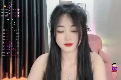 Media: A video of an East Asian woman with long black hair, fair skin, and red lipstick, sitting in a pink chair, wearing a strapless top, in a dimly lit room with black curtains.