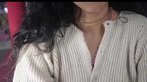 Media: Video of a person with long, wavy black hair, wearing a cream-colored, textured, V-neck sweater. The background is blurred, featuring a red cushion and indistinct, colorful objects.