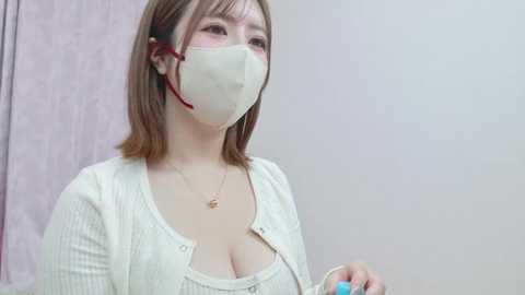 Media: Video of an East Asian woman with shoulder-length brown hair, wearing a white face mask, a white cardigan, and a low-cut top, holding a blue object, in a soft-focus indoor setting.