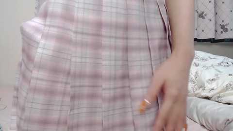 Media: Video of a person wearing a plaid, light pink and white dress, with a hand in the frame, and a bed with patterned sheets in the background.