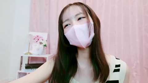 Media: Video of an East Asian woman with long, straight brown hair, wearing a pink surgical mask, a sleeveless black-and-white striped top, in a pink-draped room with a white shelf holding a potted plant.