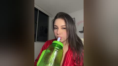 Media: A video of a young woman with light skin and long dark hair, wearing a red satin robe, drinking from a large green water bottle, indoors with a window and framed artwork in the background.