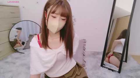 Media: Video of an Asian woman with straight brown hair, wearing a white t-shirt and beige skirt, mask, and red panties, standing in front of a mirror, reflecting her bare buttocks, in a white room.