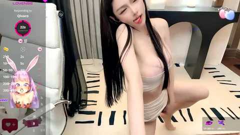 Media: Video of an Asian woman with long black hair, wearing a white bra and panties, sitting on a modern white chair, in a room with striped curtains and a pink bunny avatar on a screen.