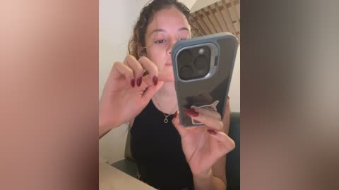 Media: Video of a young woman with curly brown hair, wearing a black top, holding a phone with a grey case, snapping a selfie. Background includes a radiator and beige wall.