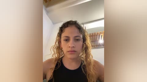 Media: Video of a young woman with light brown skin and curly, wet hair, wearing a black sports bra, standing in a white-tiled bathroom with a mirror and showerhead in the background.