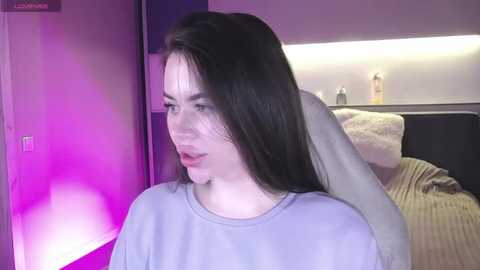 Media: Video of a young woman with long, straight black hair, wearing a light blue sweater, in a dimly lit bedroom with purple lighting, a bed with beige sheets, and a small white lamp on a nightstand.