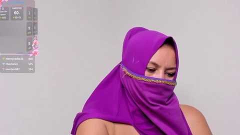 Media: Video of a woman with light skin wearing a vibrant purple hijab with golden embroidery, partially covering her face. Background features a digital screen displaying financial charts.