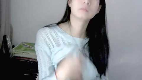 Media: Video of a young Asian woman with long black hair, wearing a light blue, see-through knit sweater that reveals her small breasts. Background shows a cluttered desk with magazines and a framed photo.