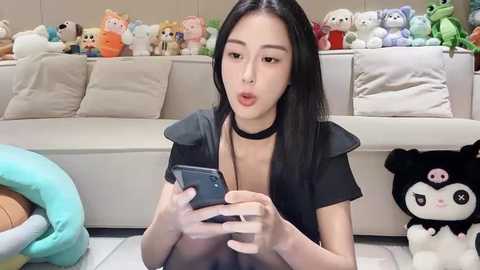 Media: Video of an East Asian woman with straight black hair, wearing a black choker and top, sitting on a white floor, playing with a smartphone, surrounded by stuffed animals on a white couch.