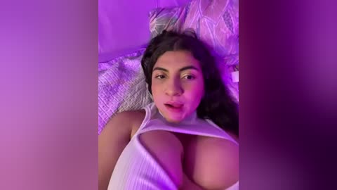 Media: Video of a Latina woman with long dark hair, wearing a white ribbed tank top that reveals her large breasts, lying on a bed with a purple light.