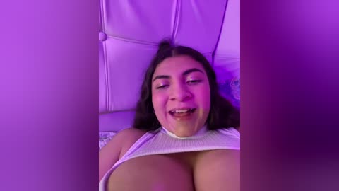 Media: Video of a smiling, light-skinned Latina woman with long dark hair, wearing a white ribbed tank top, revealing ample cleavage. She lies on a white leather couch under purple lighting.