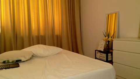 Media: Video of a minimalist bedroom with white bed, beige curtains, gold-yellow wall, white dresser, vase with yellow flowers, and mirror.