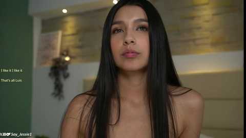 Media: A video of a young, light-skinned woman with long, straight black hair and a slender physique, wearing minimal makeup, standing in a modern, dimly-lit room with a beige wall and a textured, gold-tinged accent wall.