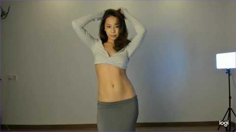 Media: Video of an East Asian woman with long brown hair, medium build, wearing a white crop top and gray skirt, posing against a plain white wall with a spotlight.