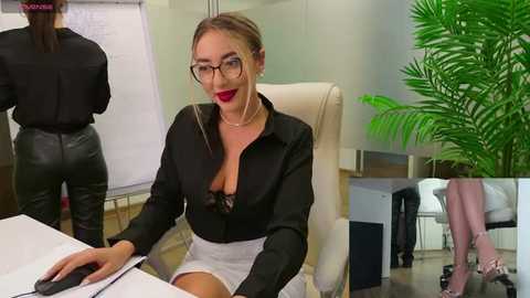 Media: Video of a seductive, glasses-wearing woman in a black blouse and gray skirt, seated at a desk, with a green plant in the background.