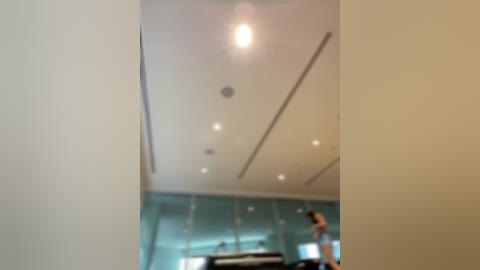 Media: Video of a modern, minimalist living room with soft, muted colors. The ceiling features recessed lighting and a white, clean design. A person in a grey shirt stands in the background, slightly blurred.