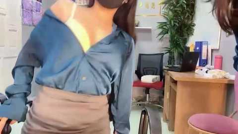 Media: Video of a woman in a blue blouse, gray skirt, and face mask, standing in a bright office with a desk, chair, and green plant.