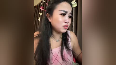 Media: Video of an Asian woman with long black hair, wearing a pink camisole, and a necklace, with a stern facial expression, indoors with brown curtains and pink ribbons.