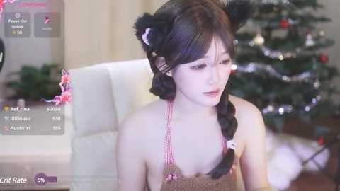 Media: Video of a young East Asian woman with long black hair in braids, wearing a brown crochet top, and black cat ears, sitting in a room with a decorated Christmas tree.