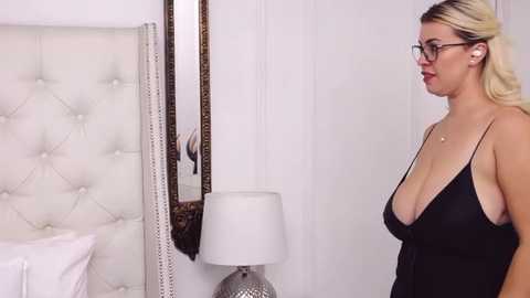 Media: Video of a blonde woman with glasses and a black dress, standing in a white and beige bedroom with a tufted headboard, a lamp, and a large mirror.