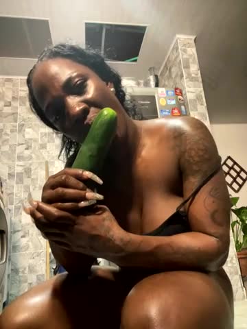 Media: Video of a dark-skinned woman with braids, holding a large cucumber, topless in a tiled bathroom with a refrigerator and colorful magnets.