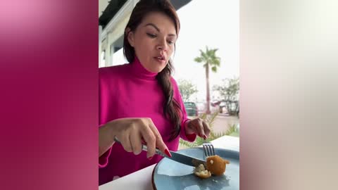 Media: Video of a woman with long brown hair in a bright pink sweater, cutting a slice of pizza with a knife and fork on a blue plate, seated outside on a sunny day with palm trees in the background.