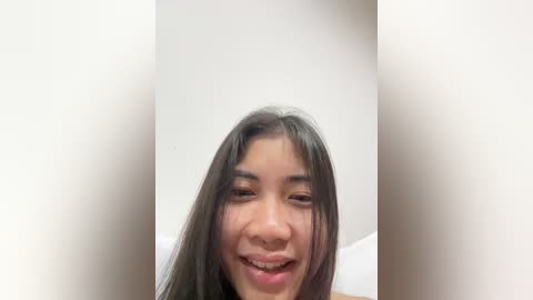 Media: Video of a smiling East Asian woman with long black hair, fair skin, and a small nose, in a white room with blurred background.