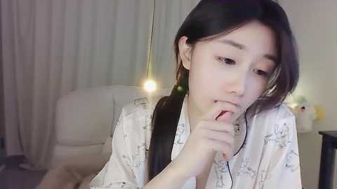 Media: Video of an Asian woman with long black hair, wearing a white robe with light floral patterns, thoughtfully biting her finger, in a dimly-lit, modern living room with white curtains and a beige couch.