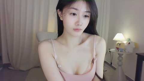 Media: Video of an East Asian woman with long black hair, wearing a pink spaghetti-strap dress, sitting indoors, with a white lamp and beige curtains in the background.