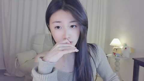 Media: Video of an East Asian woman with long black hair, wearing a gray sweater, seated indoors. She has a contemplative expression, hand near mouth. Background features a white chair, a lamp, and plush toys.