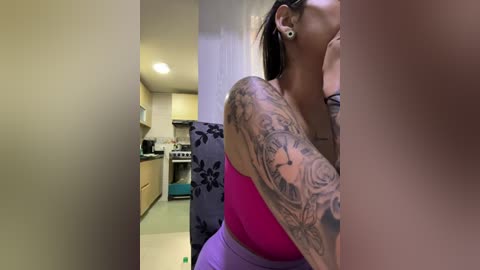 A video shows a tattooed woman in a vibrant pink top, taken from a low angle. She has a large, intricate arm tattoo. The background features a modern kitchen with light cabinets and a blue floral cushion.