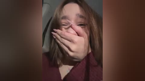 Media: Video of a woman with light skin, shoulder-length brown hair, wearing a burgundy sweater, covering her face with her hands, eyes closed, in a dimly lit room with blurred background.