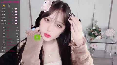 Media: Video of a young East Asian woman with long black hair, wearing a beige knitted sweater, adjusting her hair. Background features a glass table with pink flowers.