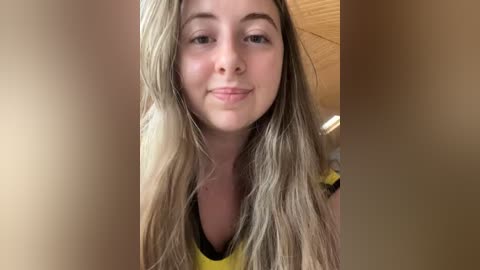 Media: Video of a young Caucasian woman with long, straight blonde hair, light skin, and a slight smile. She wears a yellow shirt with black trim. Background features a wooden wall.