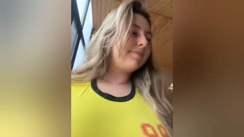 Media: Video of a smiling, light-skinned woman with long, wavy blonde hair, wearing a yellow soccer jersey with black trim and the number \"60\" in red. Background includes wooden paneling and a partially visible window.