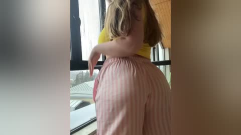 Media: Video of a curvy woman with fair skin, wearing a yellow top and pink striped pants, leaning against a window, looking outdoors.