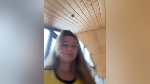 Media: Video of a young woman with long blonde hair and a yellow shirt, smiling, in a wooden-paneled room with large windows and a slanted ceiling.