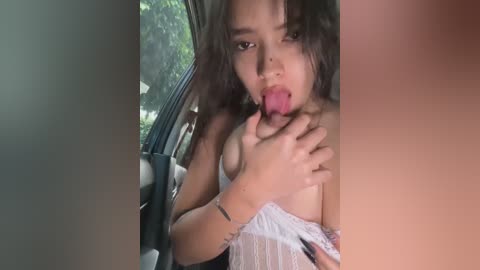 Media: Video of a young woman with long dark hair, wearing a white off-shoulder top, licking her fingers while inside a car.