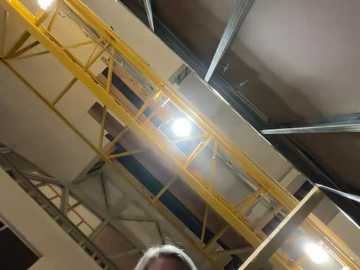 Media: Video of a partially constructed warehouse interior with exposed metal beams, yellow safety rails, and fluorescent lights. A man with gray hair stands in the foreground, partially visible.