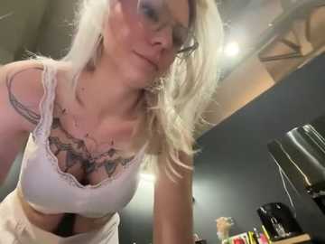 Media: Video of a blonde woman with glasses, wearing a lacy white bra, leaning over a counter. She has tattoos on her chest. Background shows kitchen items and dim lighting.