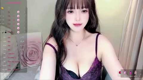 Media: Video of a young Asian woman with long black hair, wearing a purple lace bralette, sitting in a bedroom with a rose-patterned wall.