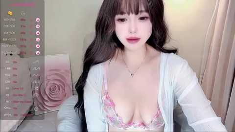 Video of a young Asian woman with long black hair, wearing a light blue cardigan over a floral-patterned bra revealing ample cleavage, sitting in a softly lit room with pastel decor.