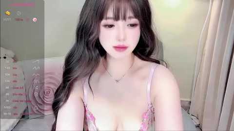 Video of a young Asian woman with long black hair, wearing a light pink lace bra, in a softly lit bedroom with a teddy bear and pastel decor.