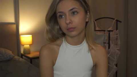 Video of a young, slender Caucasian woman with light skin and shoulder-length blonde hair, wearing a sleeveless white top, standing in a dimly lit bedroom with a warm-toned lamp, a bed, and a hanging garment.